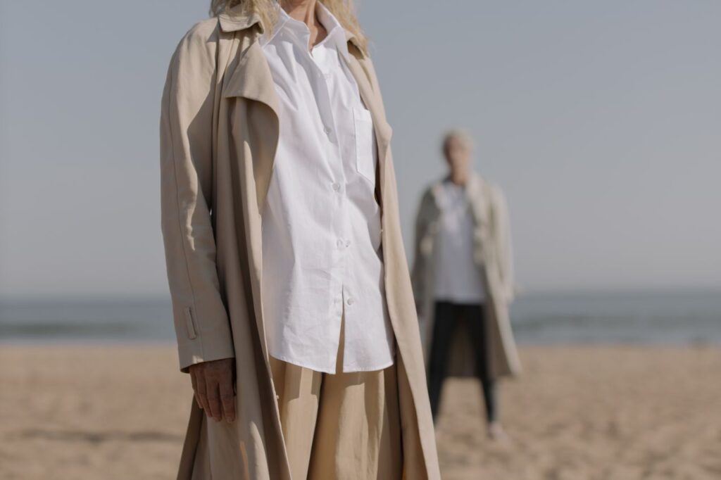 Fashionable women in beige overcoats stand on a sandy beach, showcasing elegance.